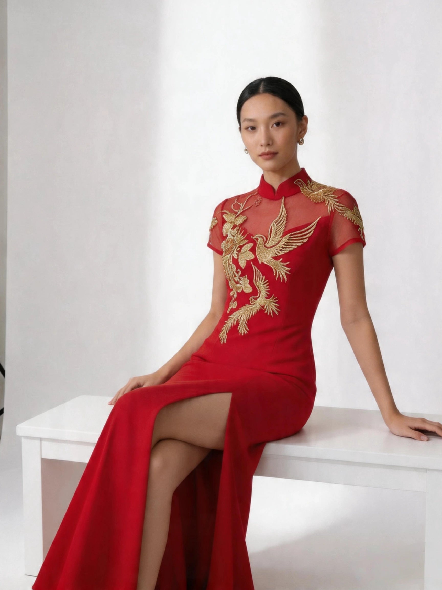 Woman wearing a Red and Golden Bird & Peony Mermaid Qipao-Modern Chinese Wedding Tea Ceremony Gown  sitting on a white bench against a white background