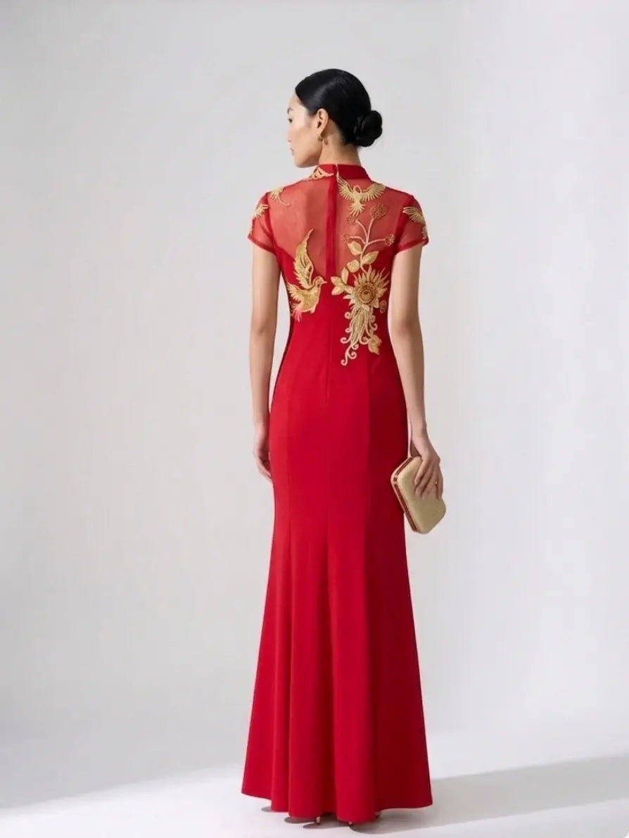 A woman wearing a red floor-length cheongsam dress with gold floral embroidery on the back, shown from behind against a light gray studio background. She is holding a small beige clutch.