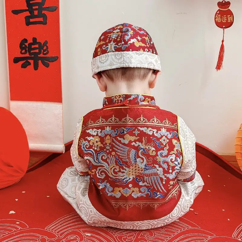 Child wearing a traditional red and gold outfit with intricate designs, sitting on a red mat.