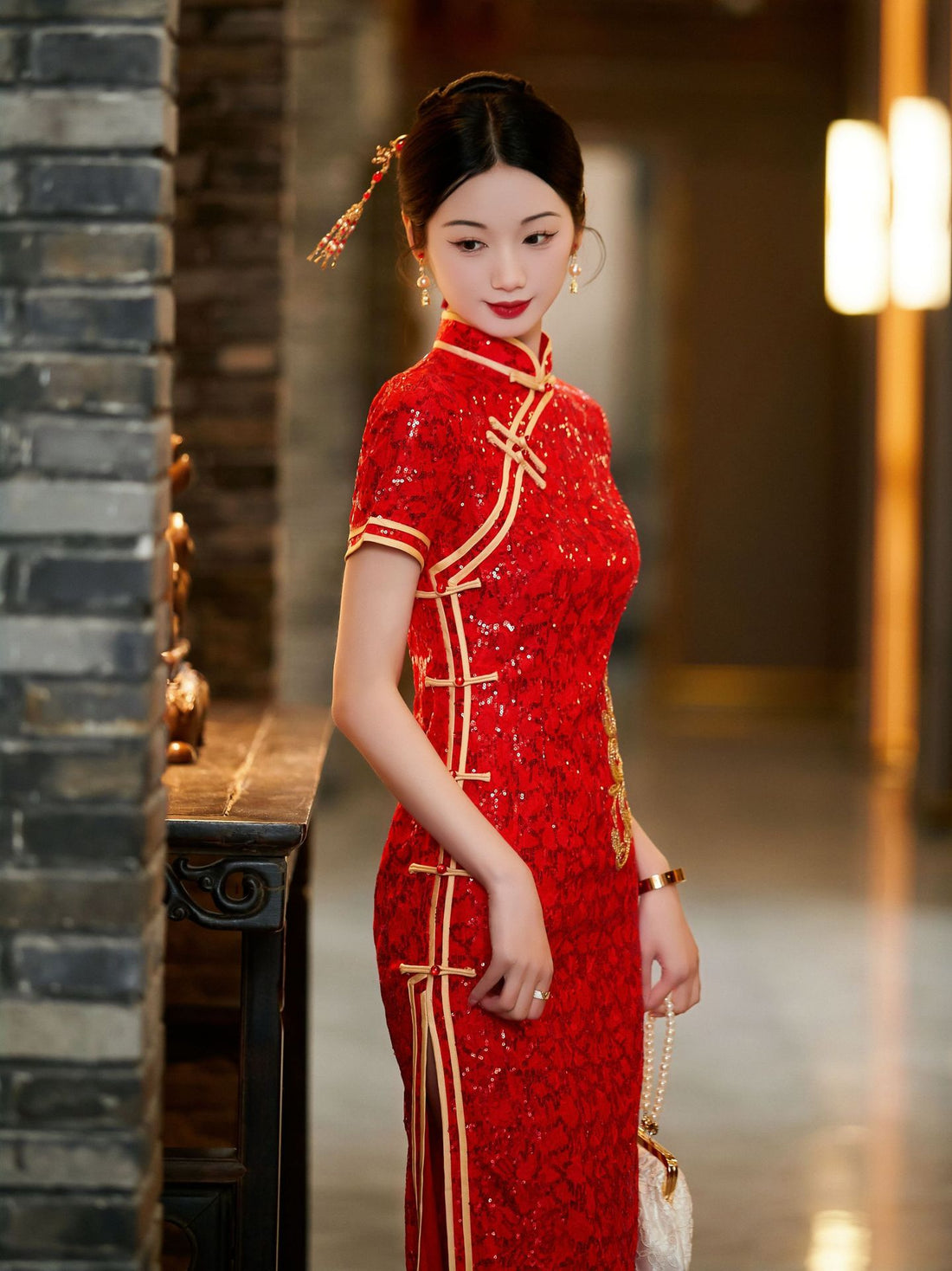 A woman wearing a red sequin qipao wedding dress with gold flower, featuring a high mandarin collar and a fitted silhouette. She holds a white satin clutch and is standing in a well-lit, elegant hallway, looking down