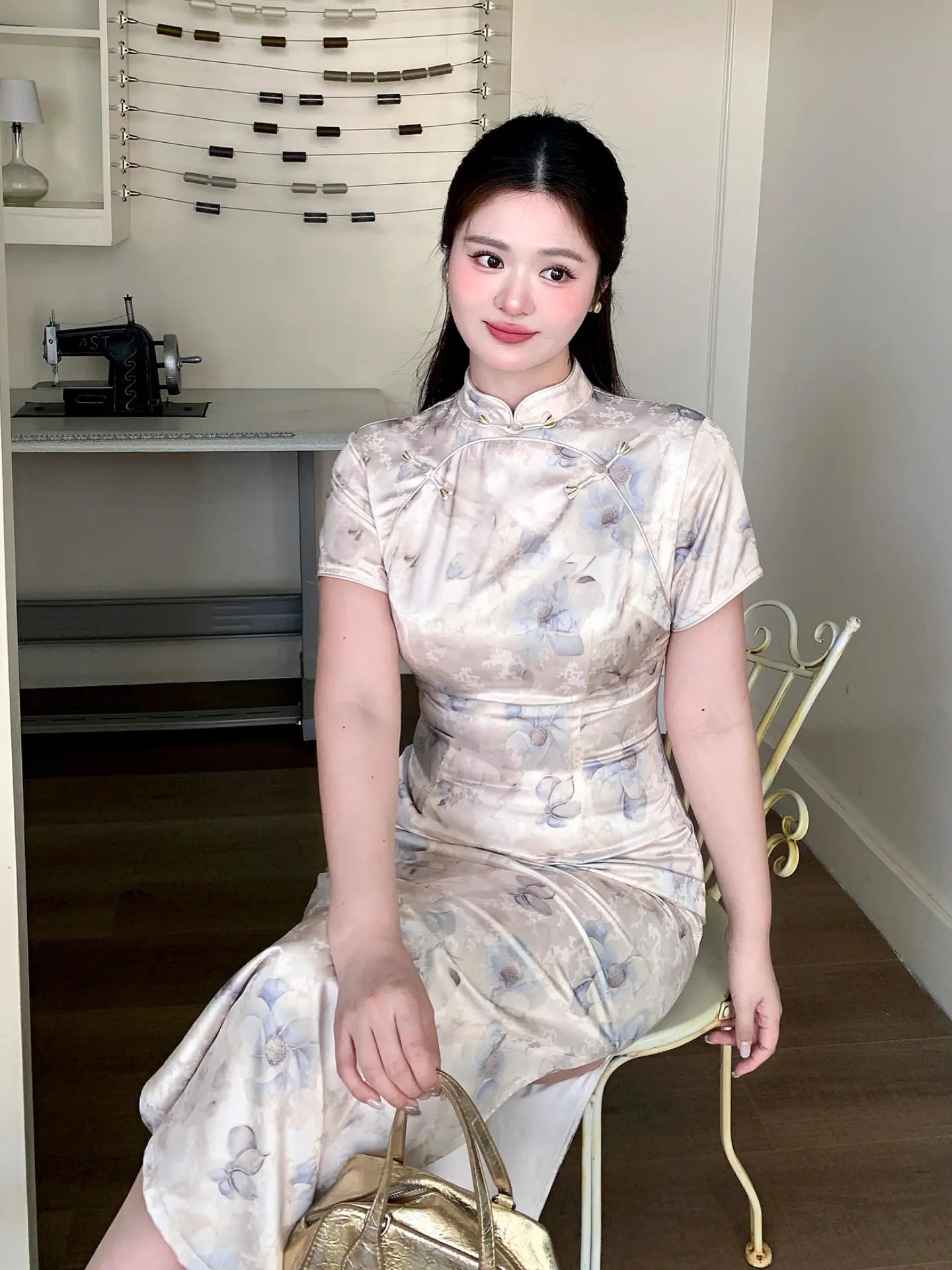 A woman wearing a plus size short-sleeved beige and gray floral print cheongsam dress, sitting on a white chair in an indoor room.