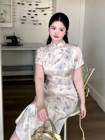 A woman wearing a plus size short-sleeved beige and gray floral print cheongsam dress, sitting on a white chair in an indoor room.