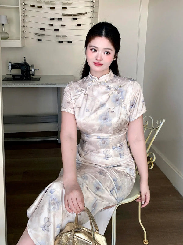 A woman wearing a plus size short-sleeved beige and gray floral print cheongsam dress, sitting on a white chair in an indoor room.