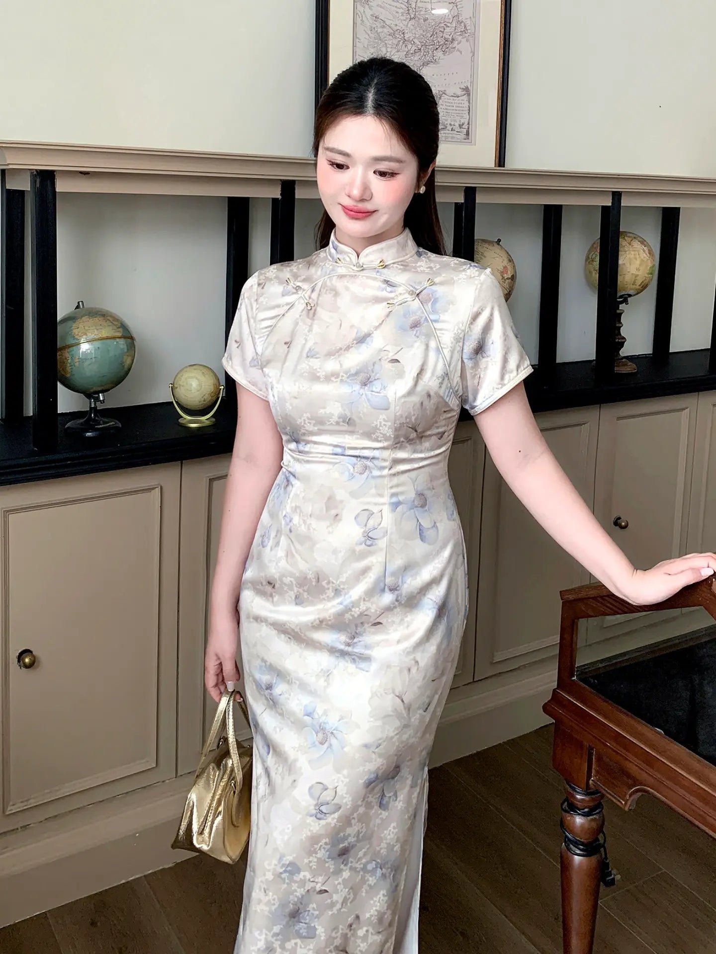 A woman wearing a short-sleeved, high-neck beige and gray floral print cheongsam dress, holding a gold handbag and posing in an indoor room with vintage globes on display.