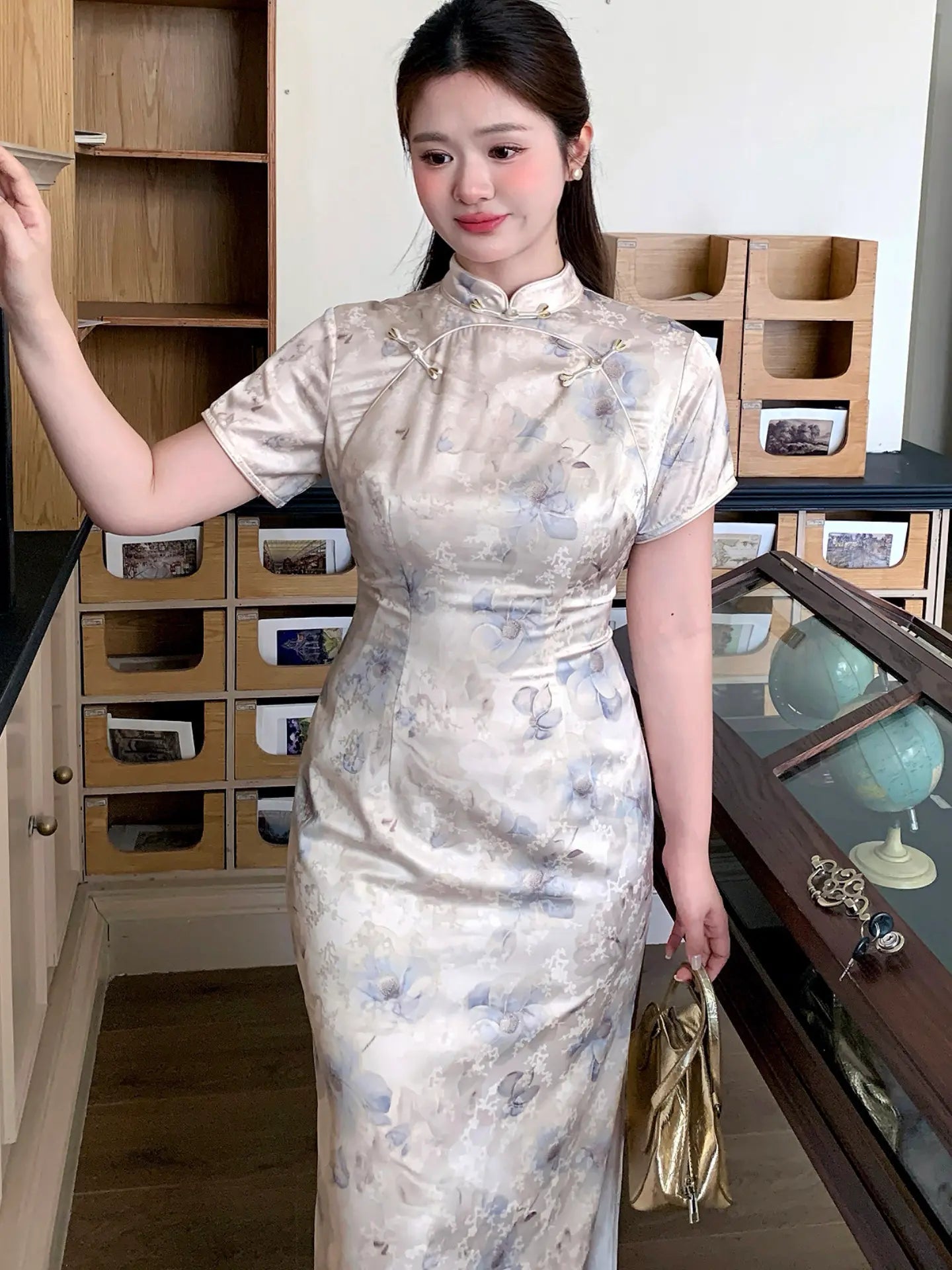 A woman wearing a short-sleeved beige and blue floral print cheongsam dress, standing in an indoor room with wooden cabinets and display shelves.