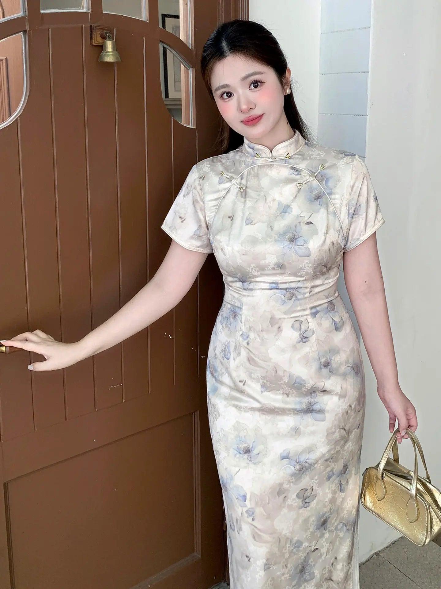 A woman wearing a short-sleeved beige and blue floral print cheongsam dress, posing in front of a brown wooden door and holding a small gold handbag.