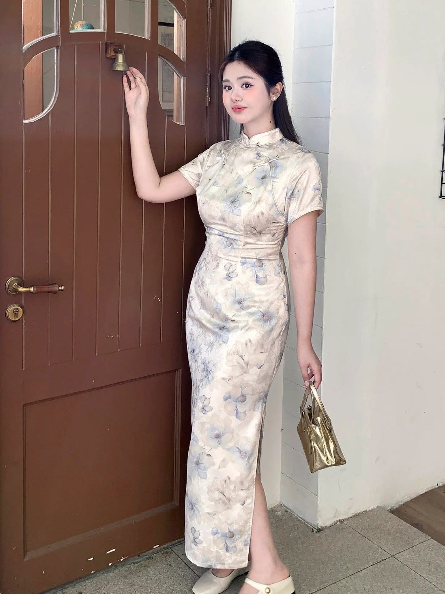 A woman wearing a beige and blue floral print cheongsam dress, posing next to a brown wooden door and holding a metallic gold handbag.