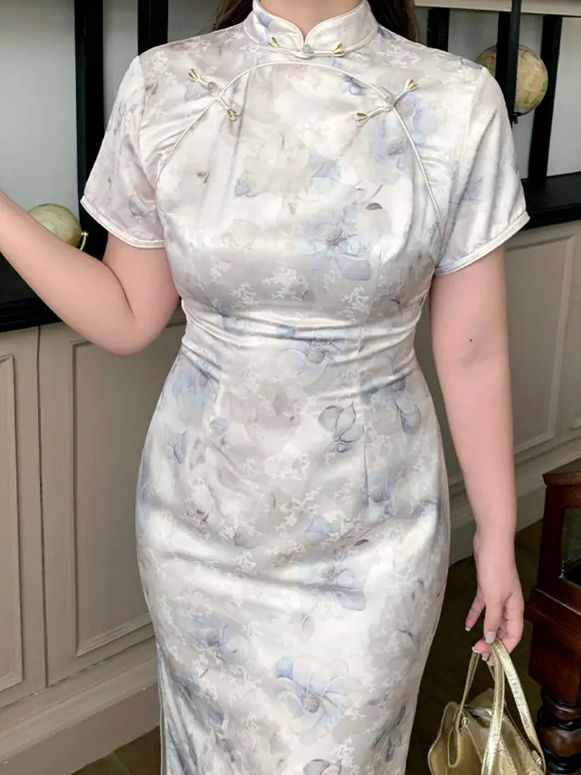 A woman wearing a short-sleeved, high-neck cheongsam-style dress with a light beige base and a soft blue floral pattern, holding a gold handbag in an indoor setting.