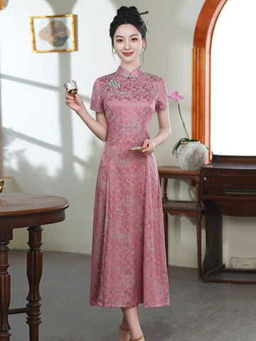 A woman stands indoors wearing a pink floral-patterned a line qipao cheongsam dress with short sleeves and a high collar. She holds a small object in one hand and stands in a styled room with wooden furniture, a window, and soft natural light.