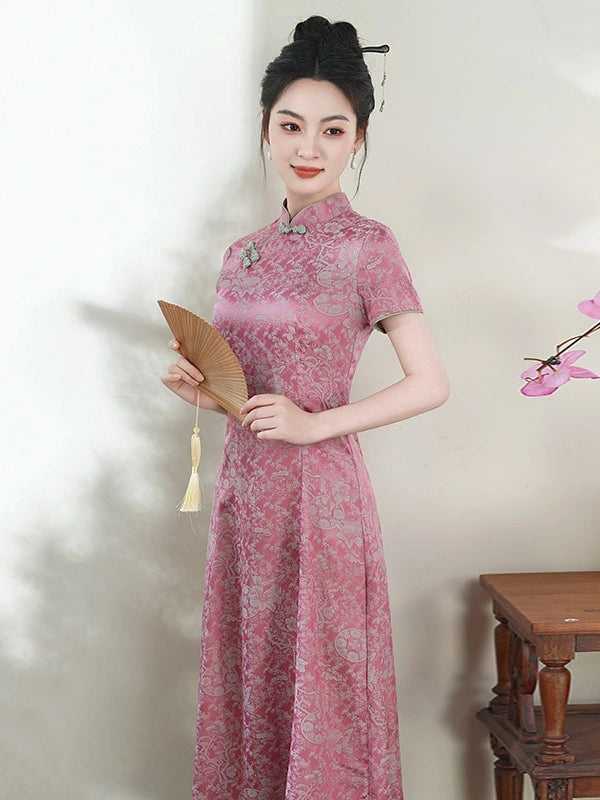 A woman wearing a pink short-sleeve cheongsam dress with a tonal floral pattern, holding a traditional folding fan. She is posed against a light beige wall with soft shadows, a wooden table at the bottom right, and a pink flower branch on the right side.
