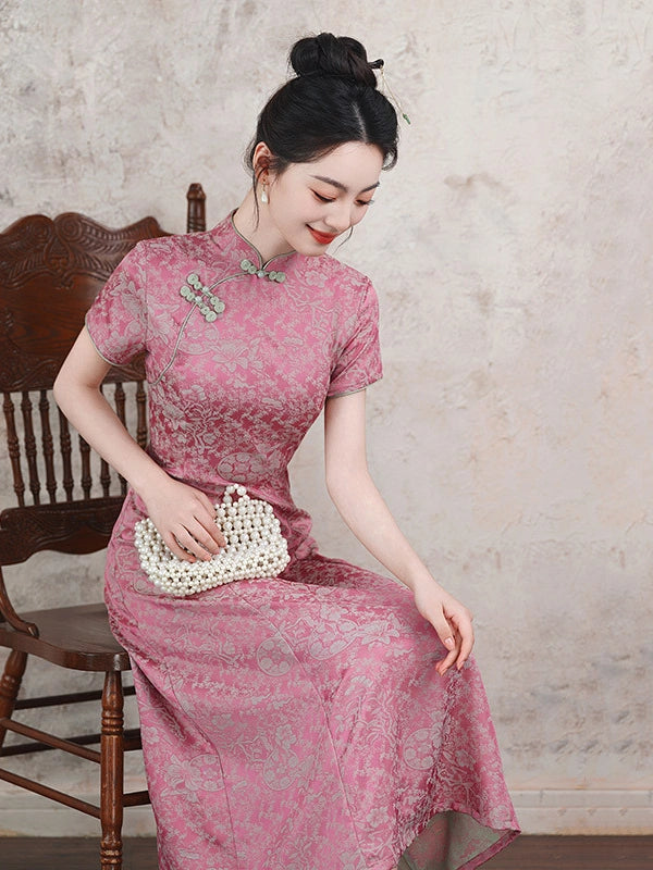 A woman wearing a pink short-sleeve qipao cheongsam dress with a subtle floral pattern, holding a white woven clutch purse while sitting on a wooden chair against a textured beige wall.