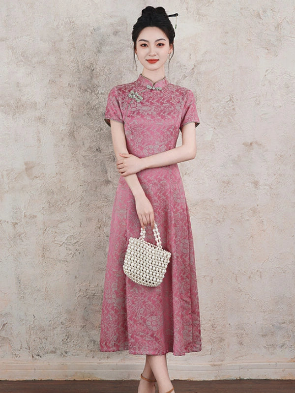 A woman models a pink short-sleeve a line qipao cheongsam dress with a subtle floral pattern, holding a white woven handbag against a textured beige wall.