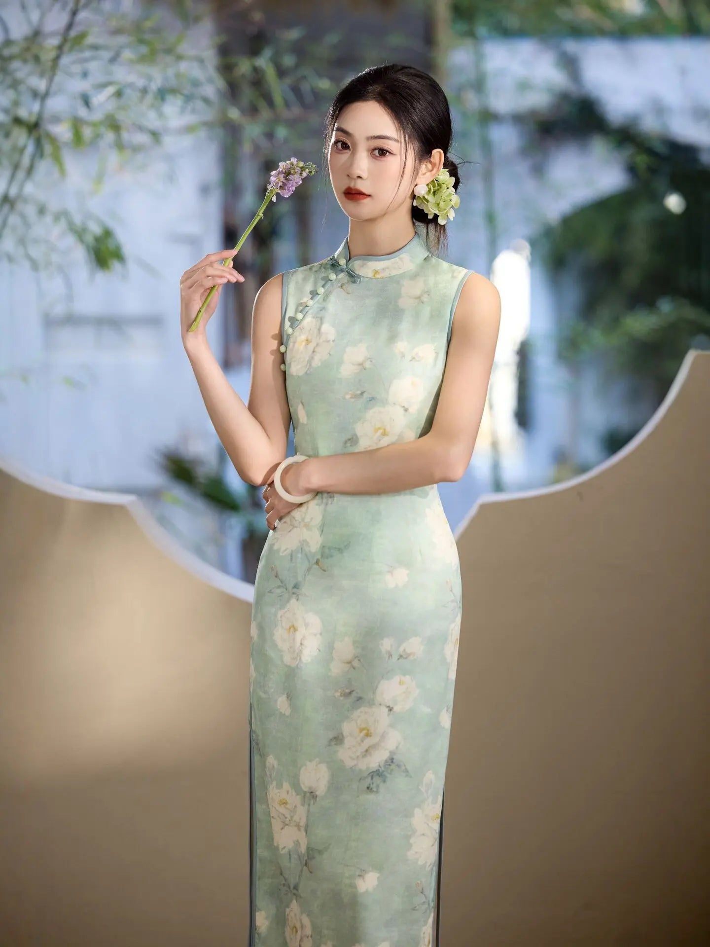 Woman in a light  Sage Green Floral Neo-Chinese Sleeveless Qipao Dress floral dress holding a flower against a blurred natural background
