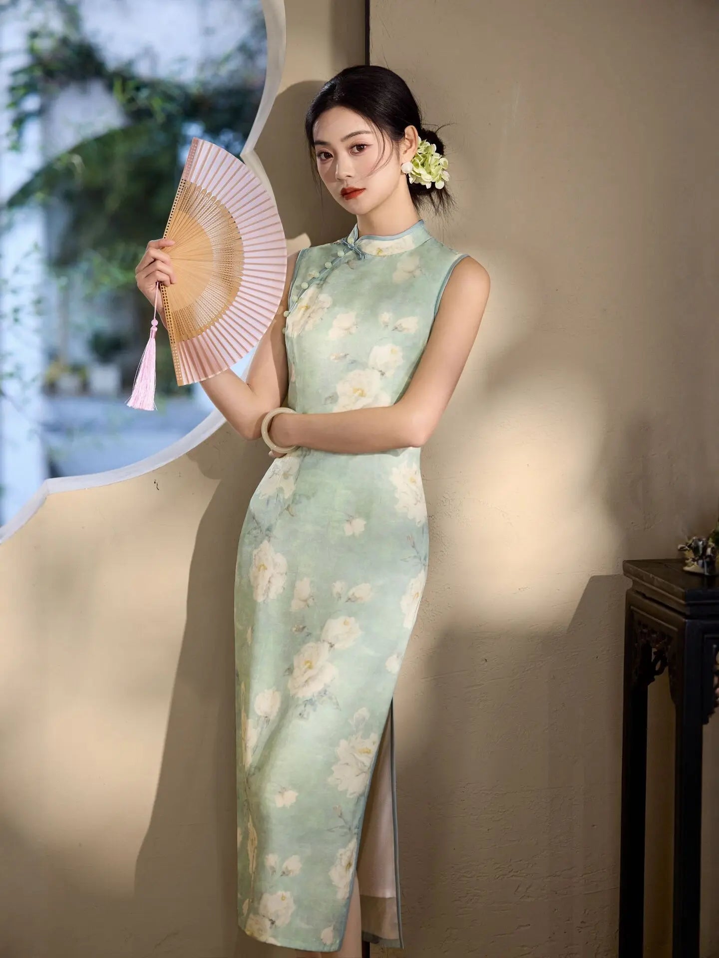 Woman in a Sage Green Floral Neo-Chinese Sleeveless Qipao Dress  holding a fan against a neutral background