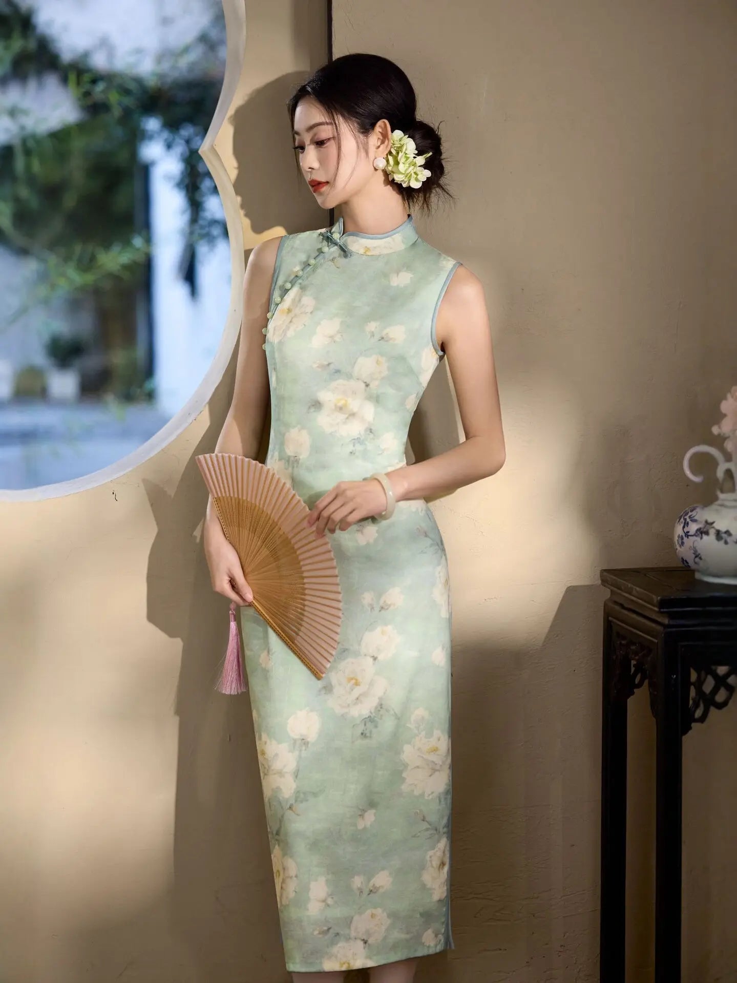 Woman in a Sage Green Floral Neo-Chinese Sleeveless Qipao Dress  holding a fan, standing in a softly lit room.
