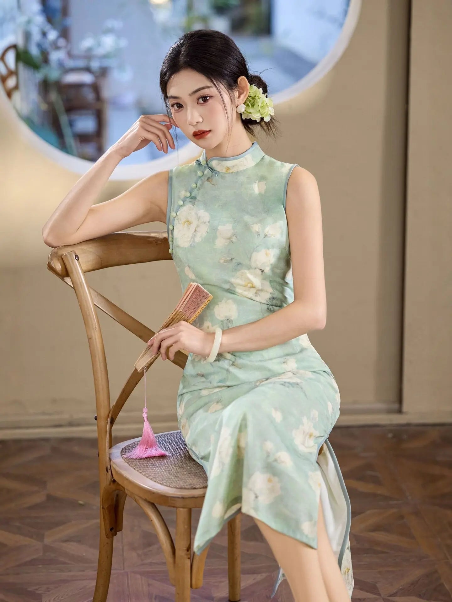 Woman in a light green Sage Green Floral Neo-Chinese Sleeveless Qipao Dress sitting on a wooden chair indoors.