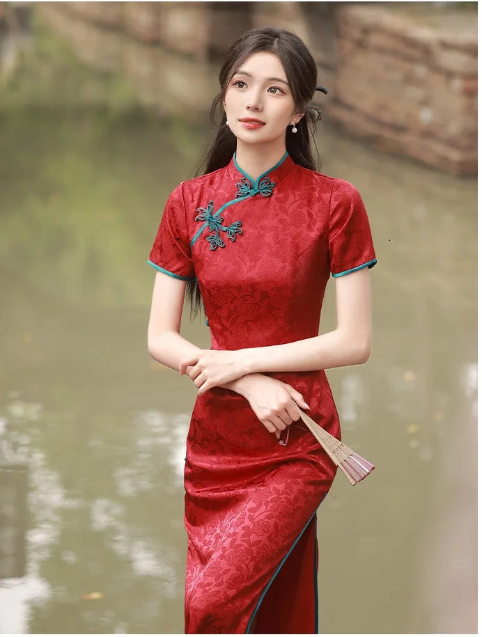Woman in a red traditional Short Sleeves Floral Jacquard Long Qipao Dress With Blue Trim  with floral embroidery standing by a body of water.