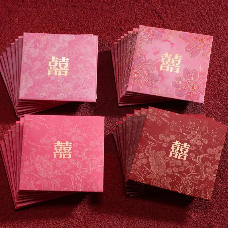 Four stacks of Chinese wedding envelopes arranged on a textured red surface. The stacks include pink and red envelopes, each printed with a gold double happiness symbol and floral patterns.