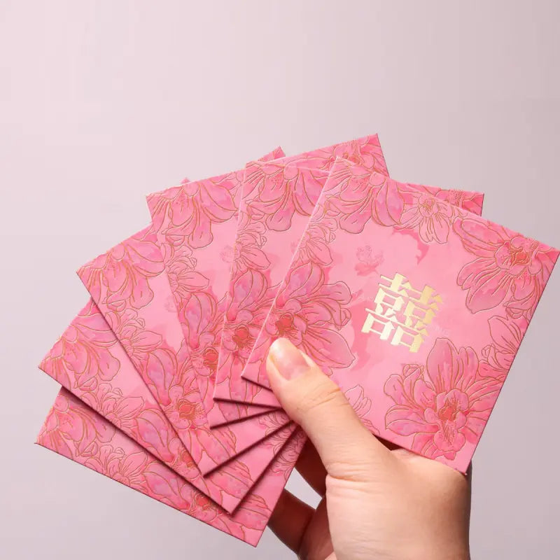 A hand holding a fan of pink Chinese wedding envelopes decorated with gold floral patterns and a gold double happiness symbol, set against a plain light gray background.