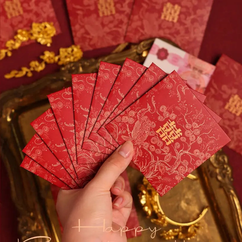 A hand holds a fan of red Chinese wedding red envelopes decorated with gold patterns and double happiness symbols, set against a festive background with gold decorations and more envelopes.