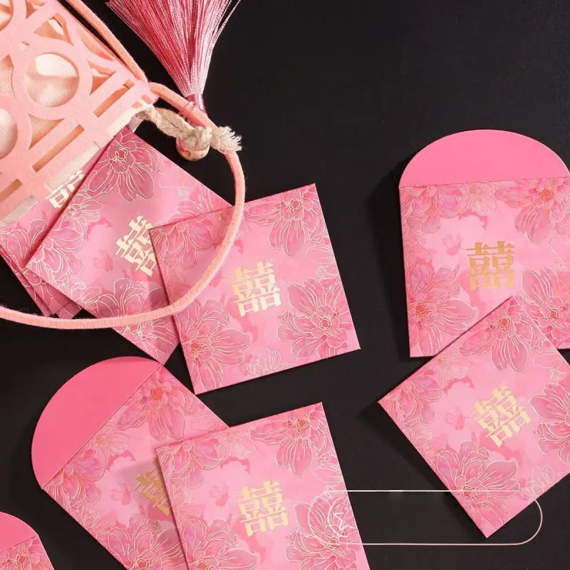 Pink floral Chinese wedding envelopes with gold double happiness symbols, arranged on a black background with a decorative pink lantern and tassels.