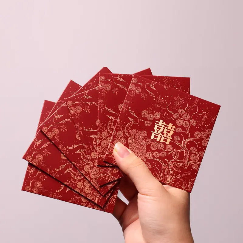 A hand holding a fan of red envelopes decorated with gold traditional patterns and Chinese double happiness symbols, set against a plain light gray background.