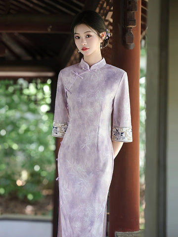 A woman wearing a Soft Lavender Jacquard  3/4 Sleeve Qipao cheongsam dress with floral embroidery and patterned cuffs, standing in a traditional wooden pavilion with green foliage in the background.