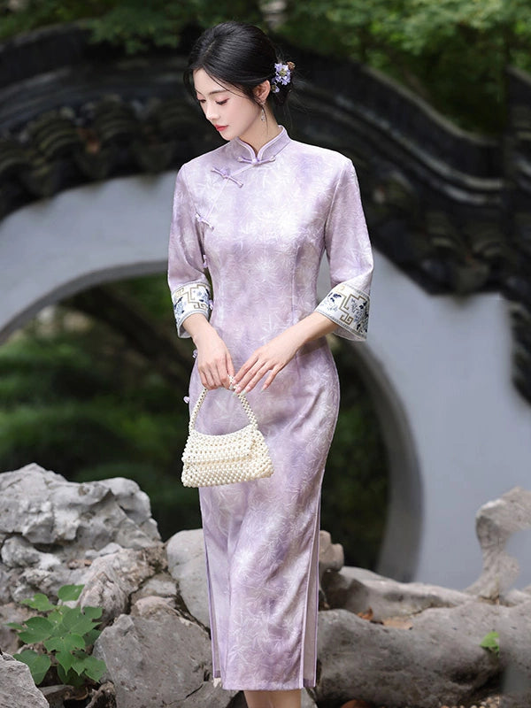 A woman wearing a light lavender-purple Chinese cheongsam (qipao) dress with patterned three-quarter sleeves, holding a small white beaded handbag in a traditional garden setting with rocks and a black tiled archway.
