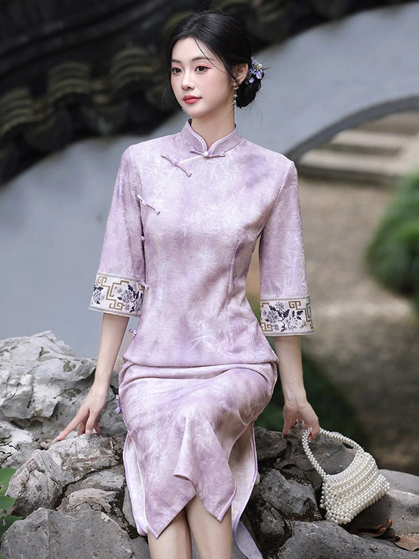 A woman wearing a light purple traditional Chinese cheongsam dress with embroidered sleeve cuffs, posing outdoors on rocks near a stone bridge.