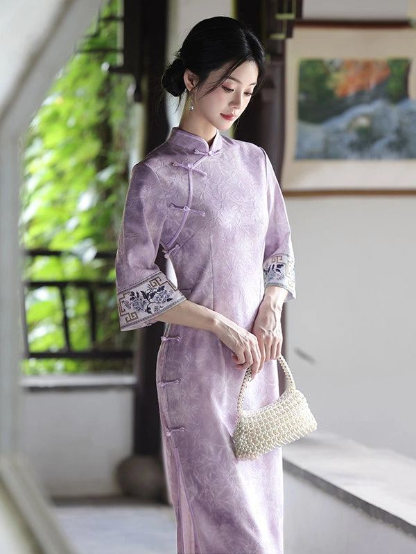 A woman wearing a lavender purple traditional Chinese cheongsam dress with embroidered cuffs, holding a small woven cream handbag in a traditional courtyard setting.