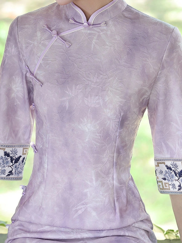 A close-up of a person wearing a lavender cheongsam-style top with a subtle tonal floral pattern, three-quarter sleeves, and decorative embroidered cuffs with blue and white floral motifs.