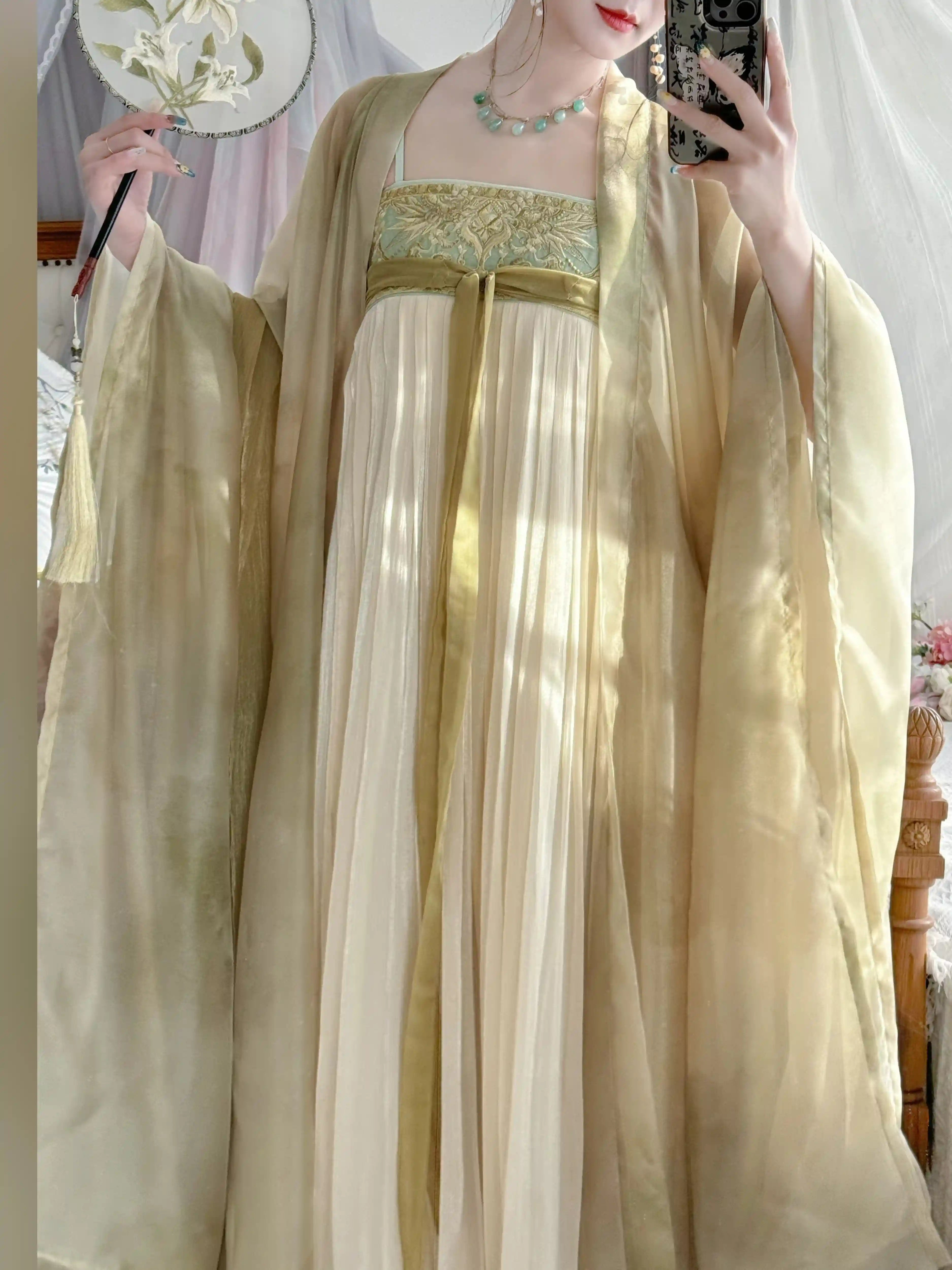 A person wearing a flowing traditional-style tang hanfu dress with wide sleeves in a soft cream and pale gold tone, holding a fan and taking a mirror selfie in an indoor setting.