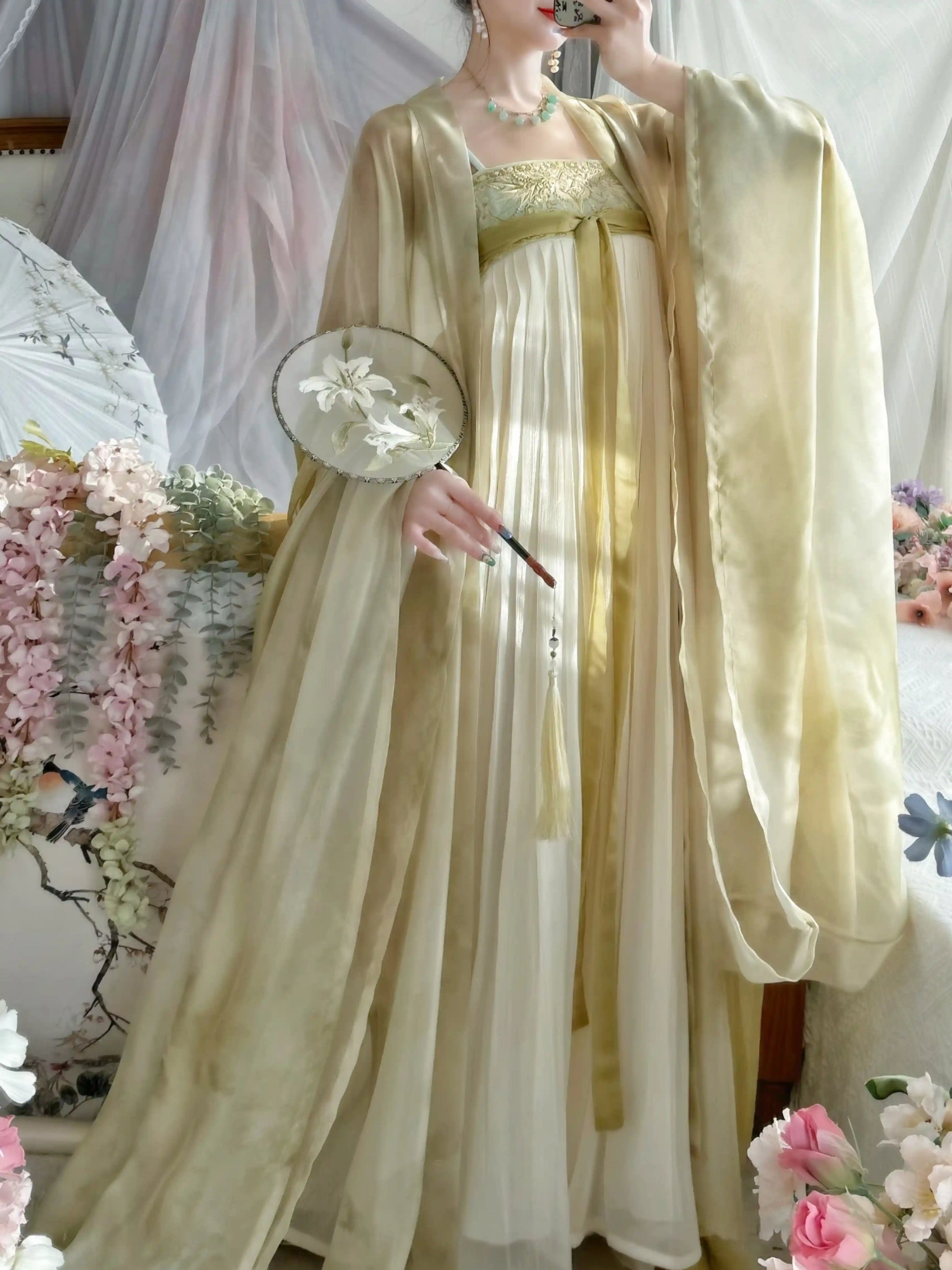 A woman wearing a flowing traditional-style dress in pale cream and soft yellow, with a matching sheer outer layer draped over her shoulders. She holds a round fan with floral embroidery, standing in a decorated indoor setting with pink flowers and draped fabric in the background.