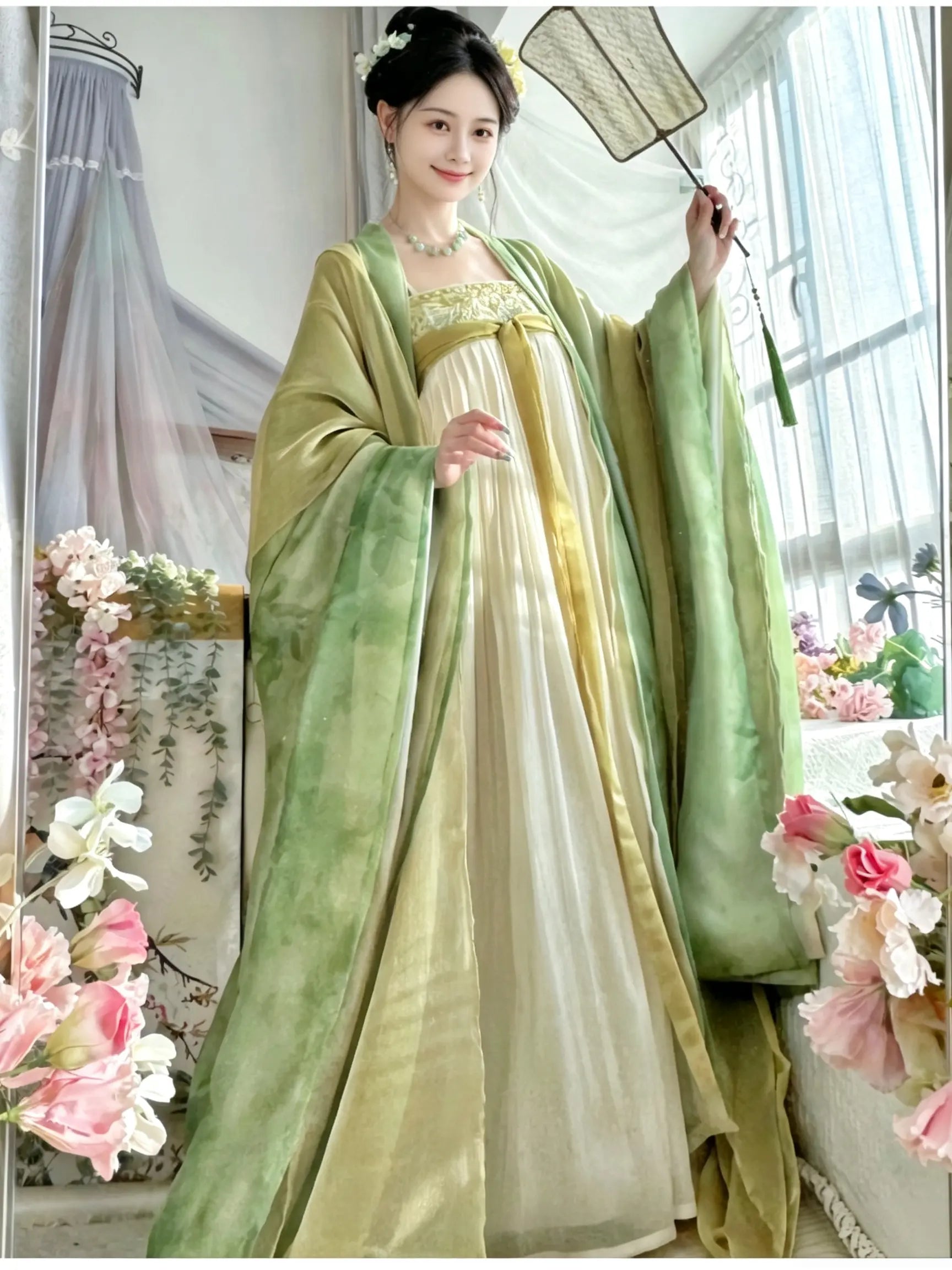 A woman wearing a traditional Chinese tang dynasty Hanfu dress in sage green and cream with gold embroidery, holding a folding fan in a decorated indoor setting.