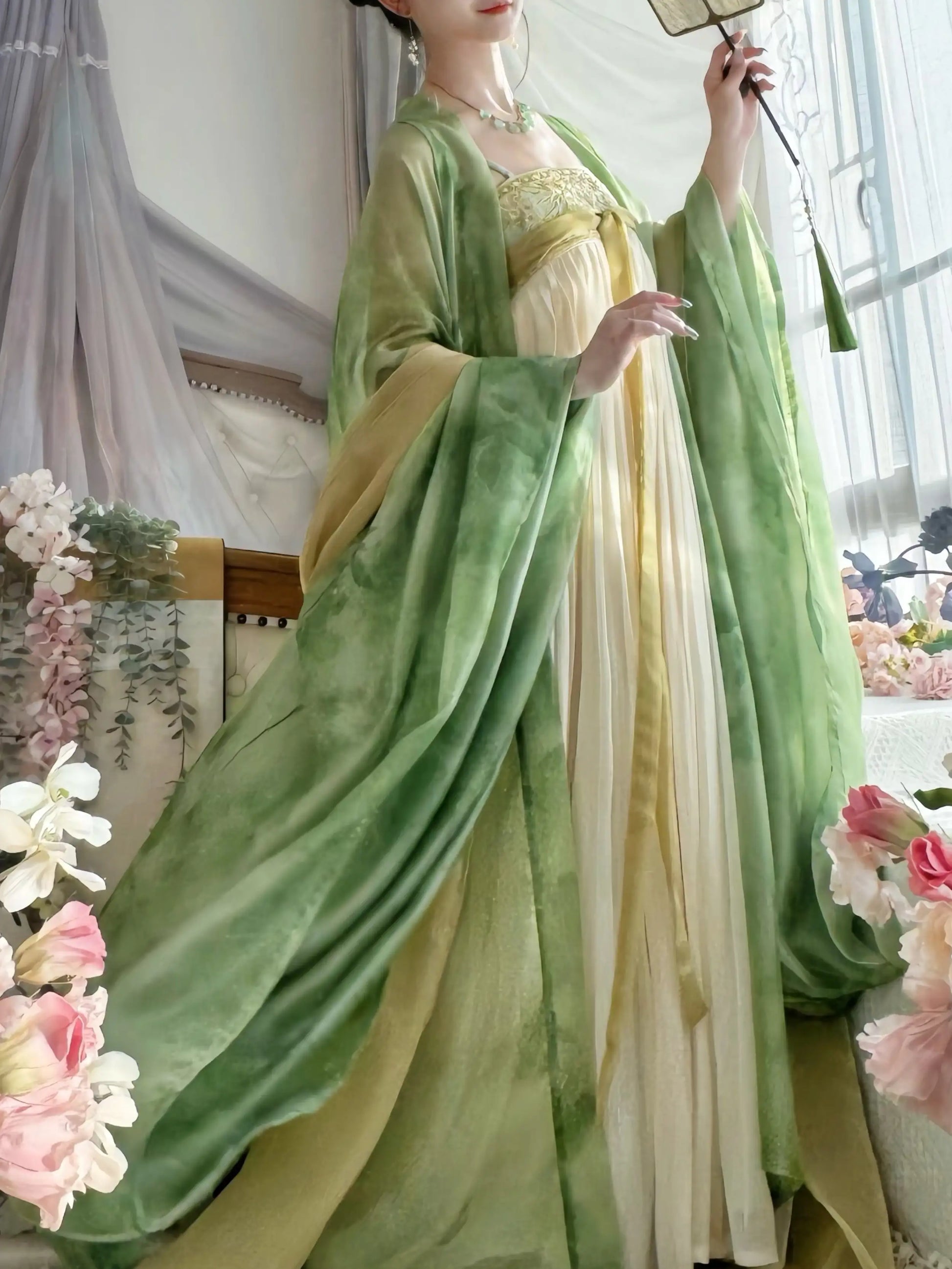 A woman wearing a flowing traditional-style tang dynasty dress with wide sleeves in shades of sage green and pale yellow, posed indoors with floral decorations and sheer curtains in the background.