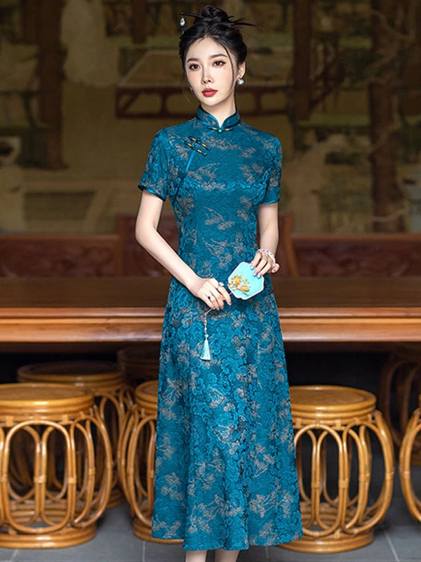 A woman stands indoors wearing a Teal Blue 3D Textured Floral Jacquard A-Line Qipao cheongsam-style dress with short sleeves and a high collar, holding a small light blue fan. The background shows wooden furniture and a softly blurred interior setting.