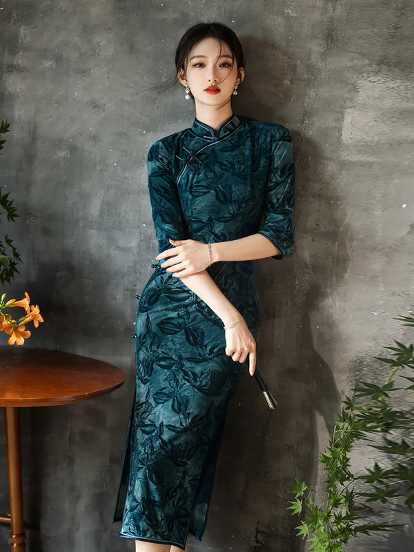 Woman wearing a Teal Foral Velvet Half Sleeves Qipao Cheongsam Dress  against a gray wall with plants.