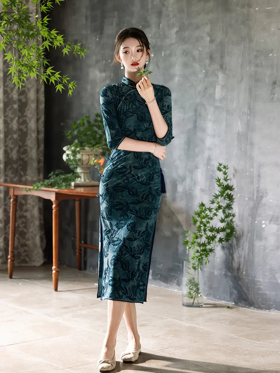 Woman in a Teal Foral Velvet Half Sleeves Qipao Cheongsam Dress  standing in a room with plants and a gray wall.