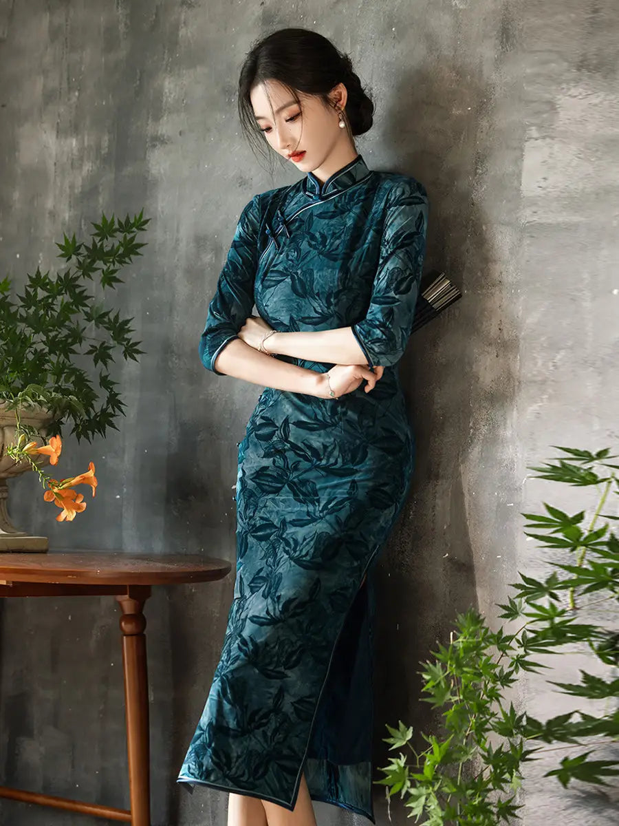Woman in a Teal Foral Velvet Half Sleeves Qipao Cheongsam Dress standing against a gray wall with plants around.