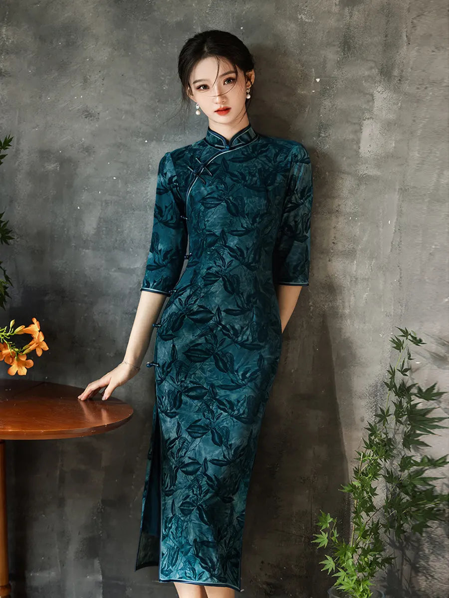 Woman in a Teal Foral Velvet Half Sleeves Qipao Cheongsam Dress standing against a gray wall with plants.