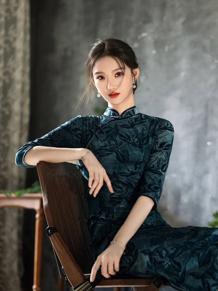 Woman in a Teal Foral Velvet Half Sleeves Qipao Cheongsam traditional outfit sitting on a wooden chair against a gray background