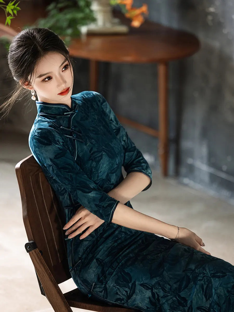 Woman in a Teal Foral Velvet Half Sleeves Qipao Cheongsam Dress  sitting on a wooden chair indoors.