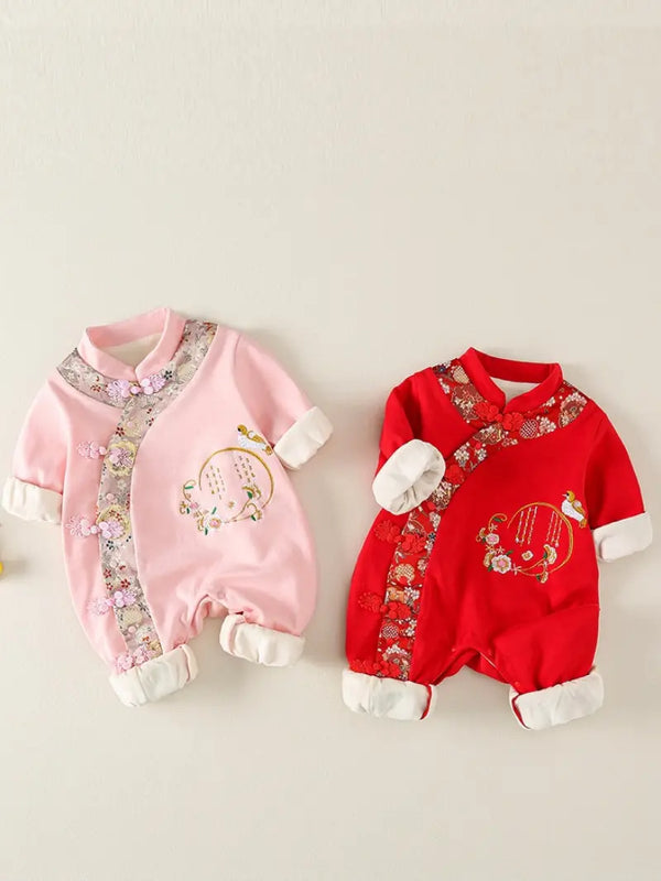 TwoTraditional Chinese Embroidered Baby Girl Qipao Romper outfits laid flat on a light beige background. One is light pink with floral embroidery and white cuffs, and the other is bright red with matching floral embroidery and white cuffs.