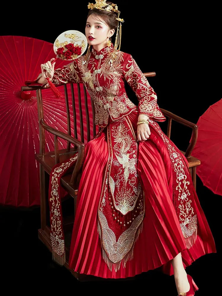 A woman seated on a wooden chair wearing an ornate red and gold traditional Chinese qun kwa xiuhe wedding dress with intricate embroidery, holding red paper fans against a black background.