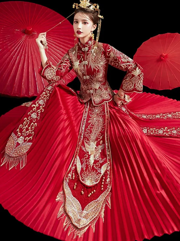 A woman wearing an ornate red traditional Chinese wedding dress with extensive gold embroidery, holding red paper umbrellas against a solid black background.