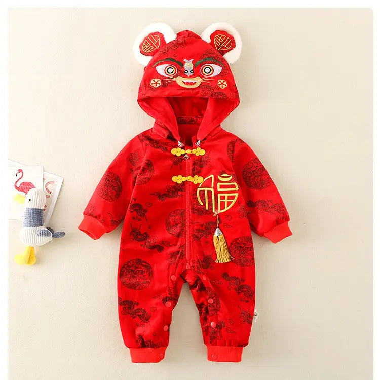 A baby's red Lunar New Year jumpsuit with a hood designed like a lion dance mask, featuring gold Chinese knot details, a gold tassel, and a gold Chinese character on the chest. The jumpsuit has a subtle black dragon pattern and is displayed against a light beige background.