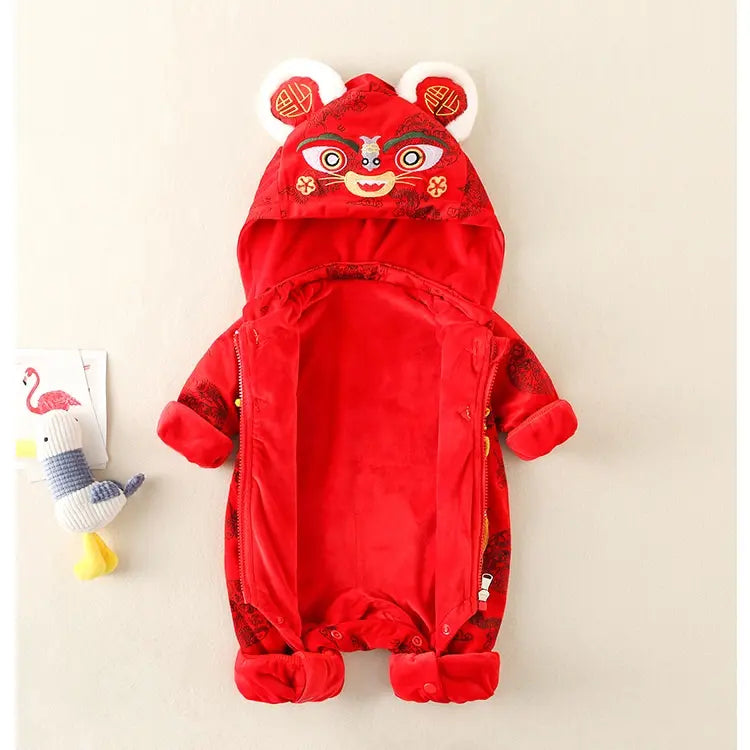 A baby's red hooded romper styled like a Chinese New Year lion dance costume, featuring embroidered facial details on the hood, plush animal ears, and a zipper front, shown on a light beige background with a small plush toy and card prop.