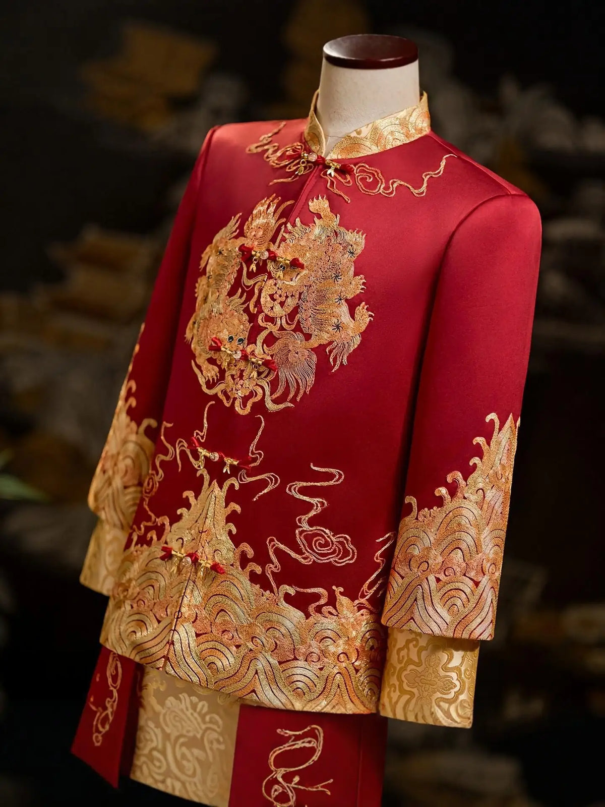 A red traditional Chinese groom wedding jacket with intricate gold dragon embroidery and gold patterned borders, displayed on a mannequin torso against a dark, blurred background.