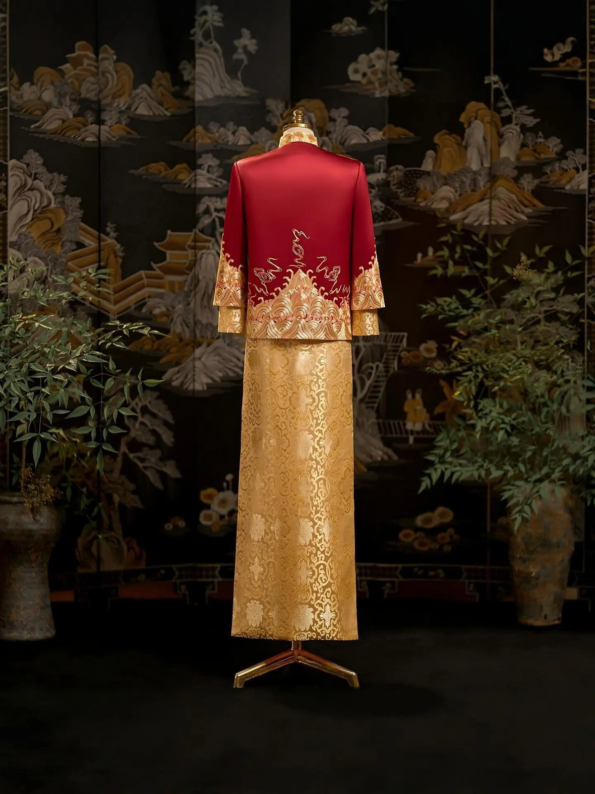 A traditional Chinese-style groom formal wedding gown displayed on a mannequin, featuring a deep red jacket with gold embroidery and a matching gold patterned skirt, set against a dark decorative screen with plants.