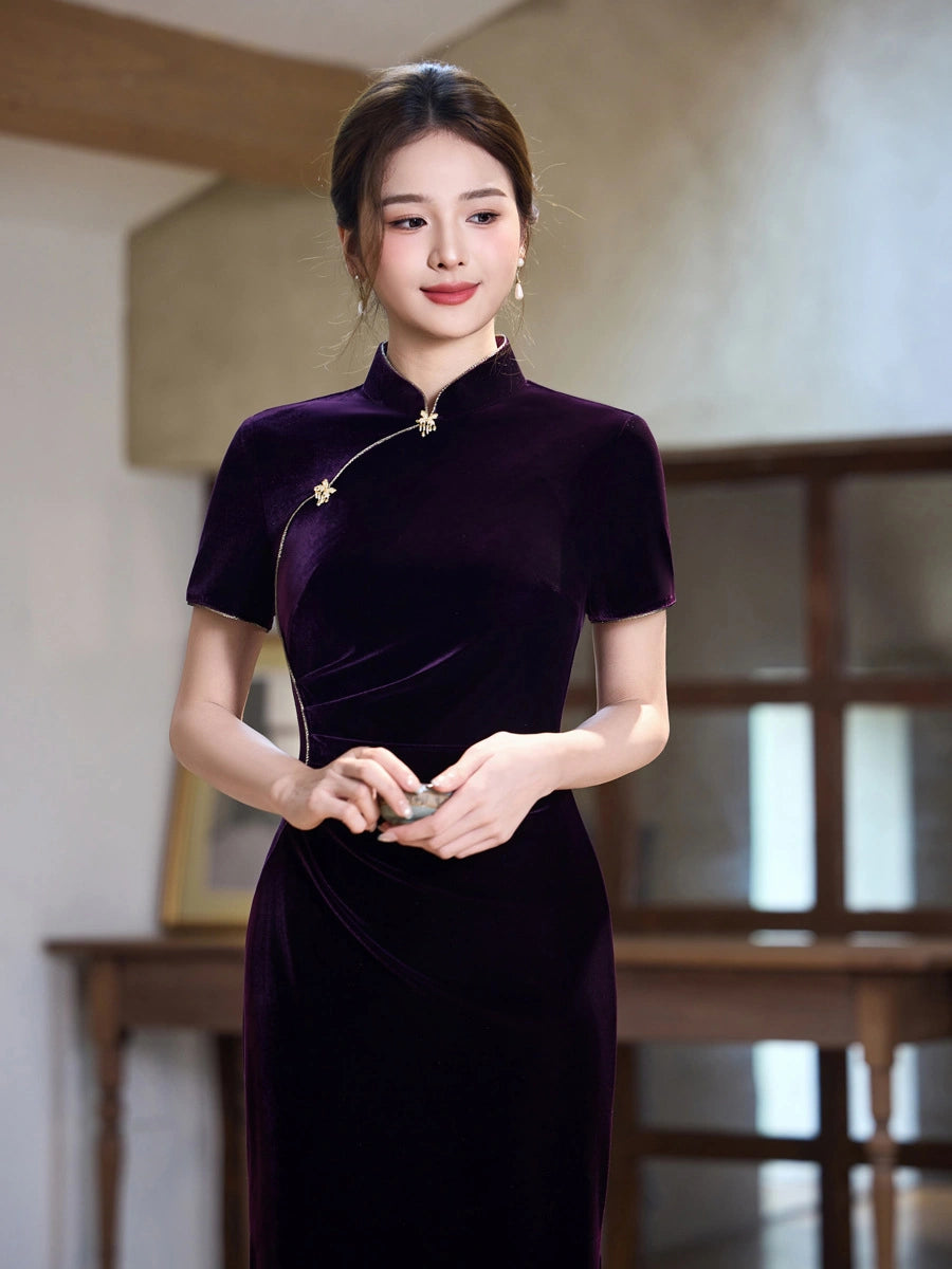A woman wearing a short-sleeved, high-neck velvet cheongsam dress in deep purple, standing in a softly lit indoor setting with a blurred wooden table and wall in the background.